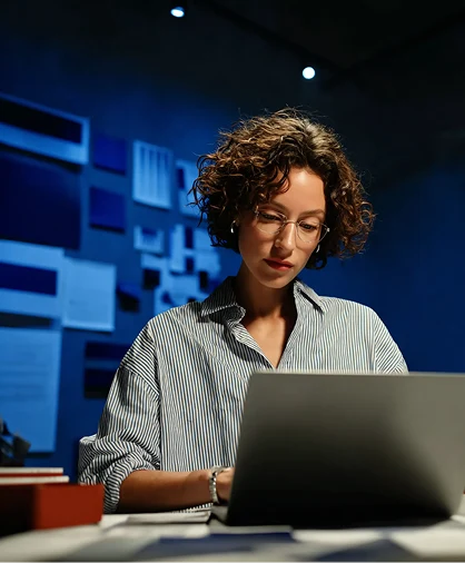 Mulher usando óculos trabalha sozinha em um notebook, em frente a uma parede com elementos visuais de identidade visual e branding
